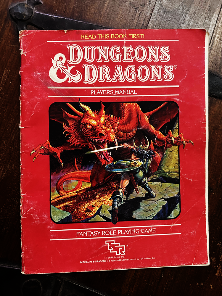 A battered old copy of the Players Manual from the 1981 Dungeons & Dragon Basic Set. It is mostly red, and features very well-painted, intensly 1980s fantasy art of a lone warrior rushing into battle against a giant red dragon.